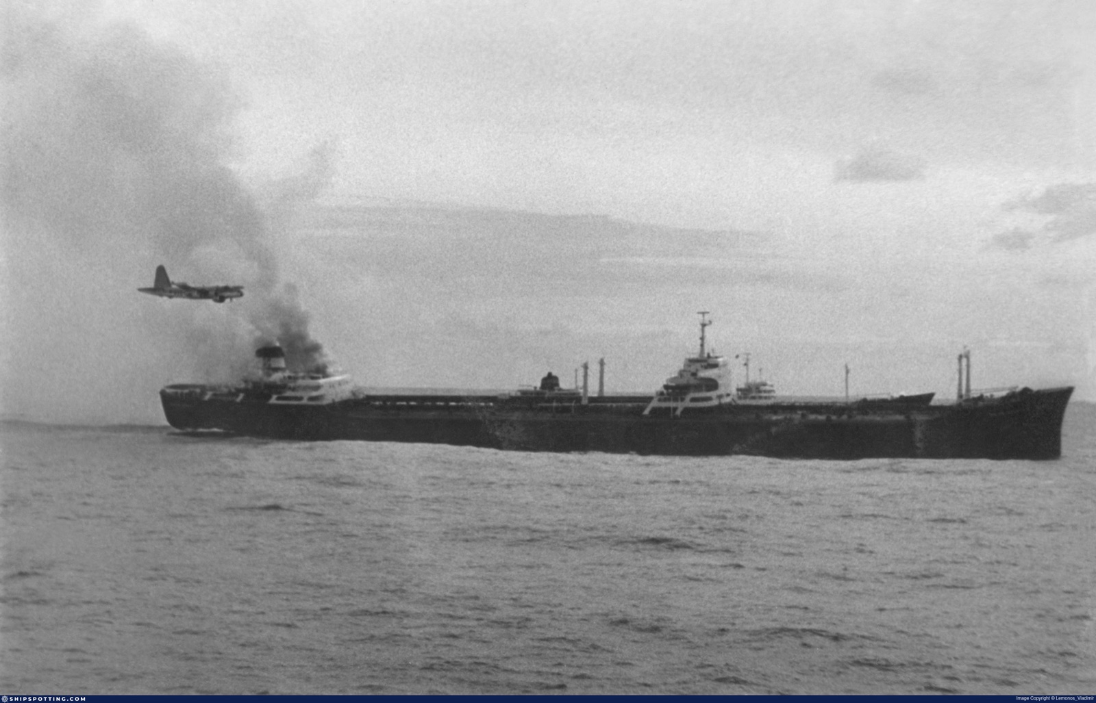 The Naess Spirit making heavy smoke, with an aircraft passing overhead.